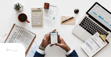 A Person Using a Smartphone Surrounded by a Laptop, and Business Documents on a White Desk